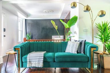 Teal velvet sofa in a bright modern living room with a brass three-arm floor lamp, patterned pillows and throw, indoor plants, dark accent wall, and hardwood floors.
