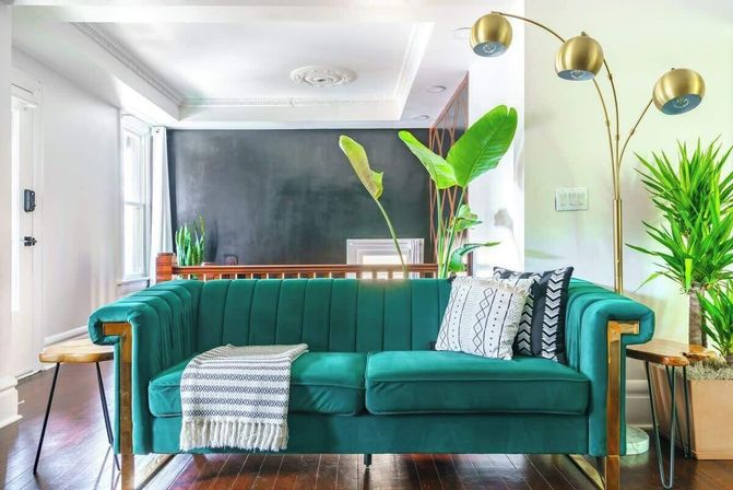 Teal velvet sofa in a bright modern living room with a brass three-arm floor lamp, patterned pillows and throw, indoor plants, dark accent wall, and hardwood floors.
