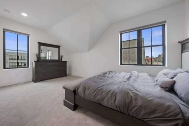 Spacious modern urban bedroom with vaulted white ceiling, two black-framed windows overlooking nearby rooftops, dark wood bed and matching dresser, gray bedding and light carpeted floor.