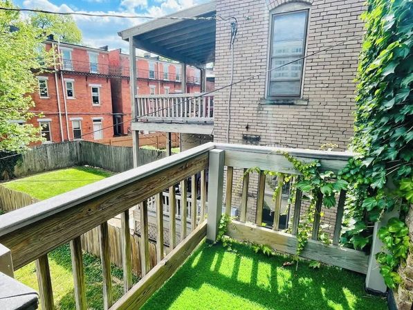 Small urban second-floor balcony with green artificial turf and wooden railing entwined with ivy, overlooking a fenced backyard and neighboring red-brick rowhouses under a sunny blue sky.
