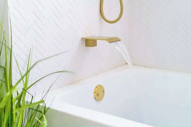 Sleek white bathtub with a gold waterfall wall-mounted faucet flowing against white herringbone tile, accented by a round gold overflow plate and green plant.