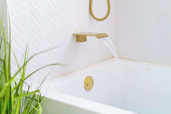 Sleek white bathtub with a gold waterfall wall-mounted faucet flowing against white herringbone tile, accented by a round gold overflow plate and green plant.