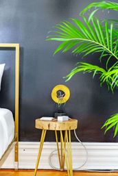Modern bedroom corner with black accent wall, gold metal bedframe, natural wood stump side table topped by a yellow circular lamp and chargers, and vibrant indoor palm fronds.