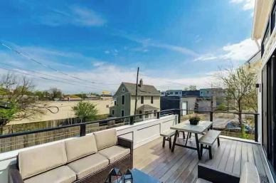 Sunny urban rooftop deck with beige wicker sofa, wooden picnic table and benches, and glass railing overlooking neighboring houses and trees under a bright blue sky.
