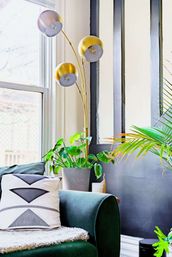 Sunlit living room corner with green velvet sofa, geometric throw pillow, potted tropical plants, and a three-globe gold arc floor lamp by a large window.