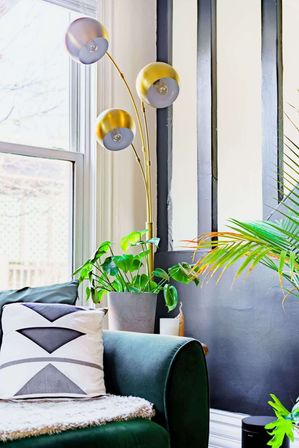 Sunlit living room corner with green velvet sofa, geometric throw pillow, potted tropical plants, and a three-globe gold arc floor lamp by a large window.