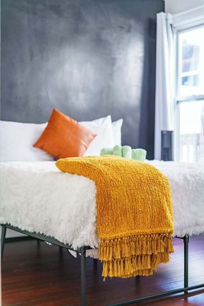 Cozy modern bedroom with white faux-fur bedding, mustard yellow fringed throw draped over the metal bed frame, orange accent pillow, gray textured accent wall and sunlit window