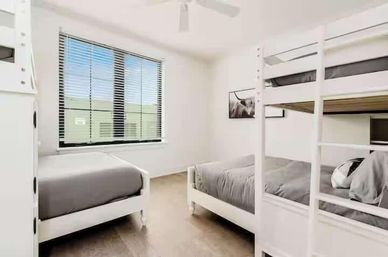 Sunlit minimalist bedroom with white bunk bed and single bed, gray linens, large window with blinds, and light wood floor.