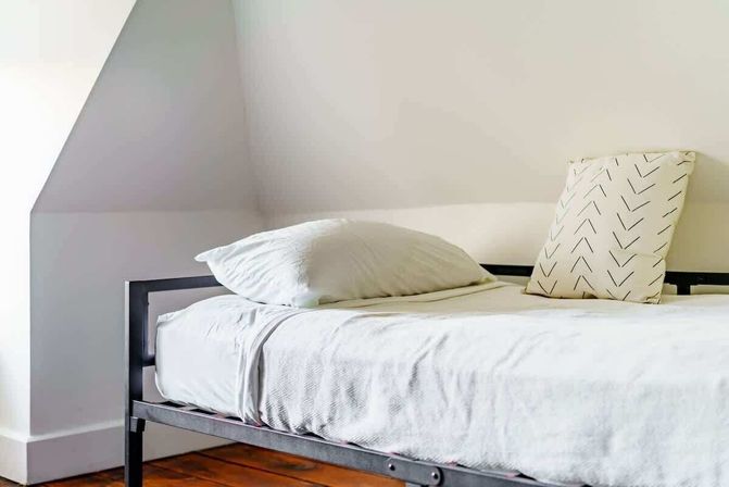 Cozy minimalist attic bedroom with white linens, arrow-pattern throw pillow on a black metal bed frame and warm hardwood floor