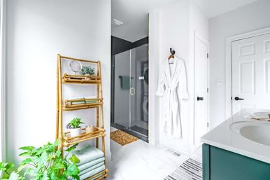 Bright modern residential bathroom with glass walk-in shower, marble tile floor, bamboo shelving holding folded mint towels and potted plants, white bathrobe on a wall hook, teal vanity and spa-like accents.