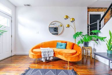 Bright modern urban living room with a curved orange velvet sofa on hardwood floors, teal accent pillow and tasseled throw, round wall mirror, gold arc floor lamp, and potted indoor plants by a white entryway.