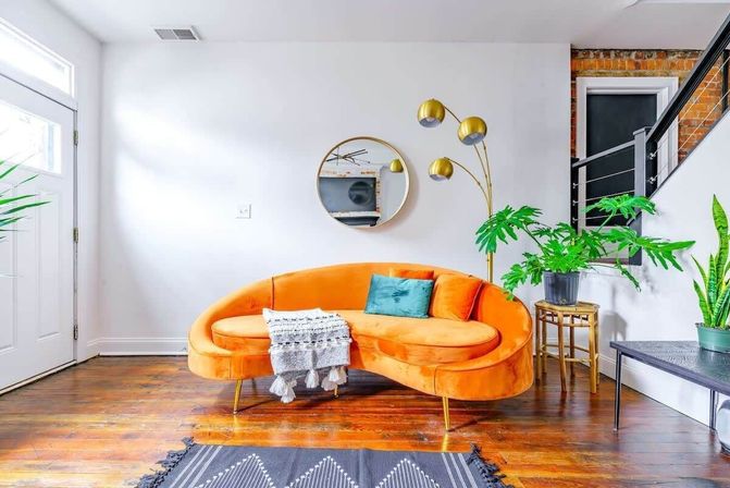 Bright modern urban living room with a curved orange velvet sofa on hardwood floors, teal accent pillow and tasseled throw, round wall mirror, gold arc floor lamp, and potted indoor plants by a white entryway.