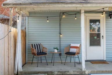 Cozy porch with pale blue siding: two rattan chairs with patterned cushions around a small round bistro table, hanging Edison-style string lights and a white door on a concrete stoop.