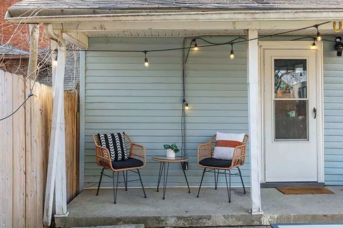 Cozy porch with pale blue siding: two rattan chairs with patterned cushions around a small round bistro table, hanging Edison-style string lights and a white door on a concrete stoop.