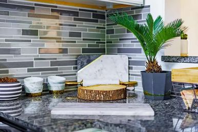 Stylish modern kitchen countertop with gray glass tile backsplash, potted sago palm, round wood-slice cutting board, marble serving boards and small ceramic bowls.