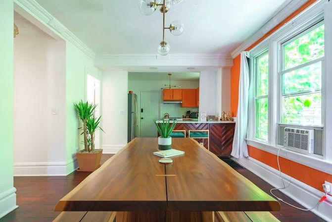 Sunlit open-plan dining room with a long live-edge wooden table, potted succulent centerpiece, orange-accent kitchen with chevron wood island, large windows and window air conditioner.