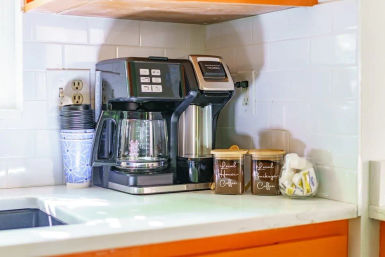 Bright kitchen coffee station on white subway-tile backsplash: black multi-function drip coffee maker with glass carafe, stack of blue patterned disposable cups, two brown coffee canisters with wooden lids, and a jar of sugar and creamer packets on a white countertop above orange cabinets, ready for morning brew.