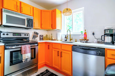 Bright modern kitchen with bold orange cabinets, stainless-steel appliances, white subway-tile backsplash, gold faucet, window light, and countertop coffee station.