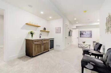 Bright, cozy finished basement lounge with a wooden wet bar and wine fridge, floating shelves and sink, carpeted neutral interior, wall-mounted TV and a small seating area with patterned chairs and coffee table.