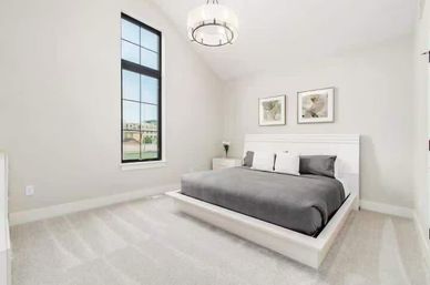 Sunlit modern minimalist bedroom with tall black-framed window, white platform king bed with gray bedding, matching nightstand, framed wall art, circular chandelier and light carpeted floor.