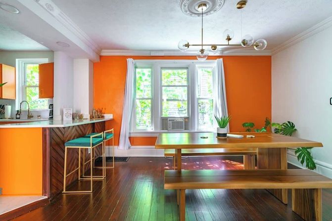 Sunlit urban apartment dining area with bright orange accent wall, wooden picnic-style table and bench, teal bar stools at a chevron wood breakfast bar, triple windows with a window AC, dark hardwood floors, brass globe chandelier and potted plants.