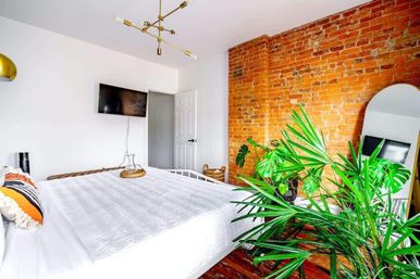 Bright urban bedroom with exposed red brick wall, white bed, large tropical indoor plants, modern gold chandelier and full-length mirror