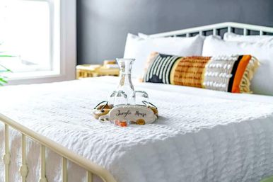 Sunlit modern bedroom with crisp white quilted bed, patterned accent pillow and a glass carafe with two glasses on a small tray at the center of the bed.