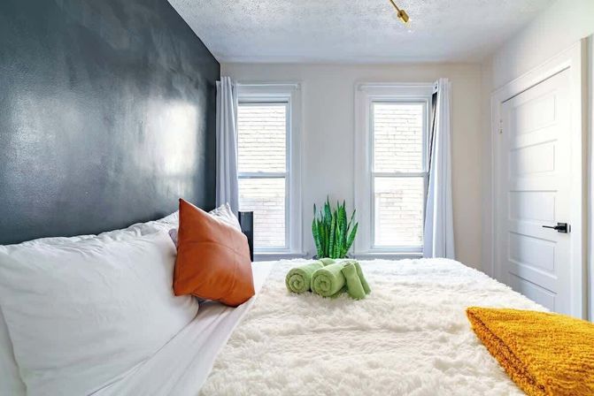 Cozy modern bedroom with white fluffy bedding, dark gray accent wall, orange throw pillow, rolled green towels and a snake plant, two tall windows with white curtains letting in natural light and a mustard throw at the foot of the bed.
