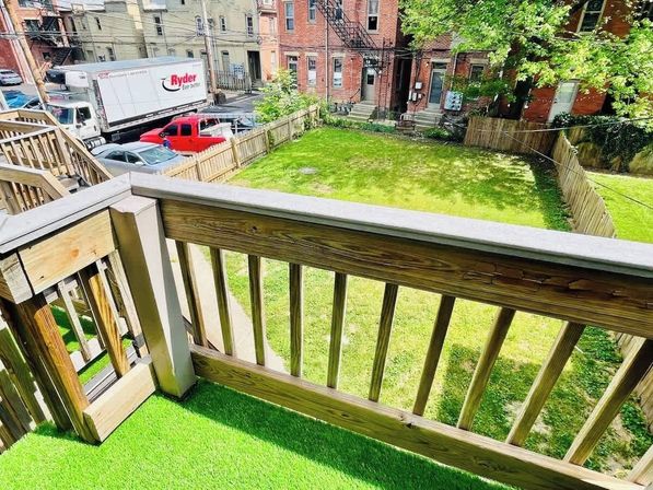 Sunny balcony view from wooden railing with green turf overlooking a fenced urban backyard lawn, parked vehicles and brick rowhouses with fire escapes in a city neighborhood.