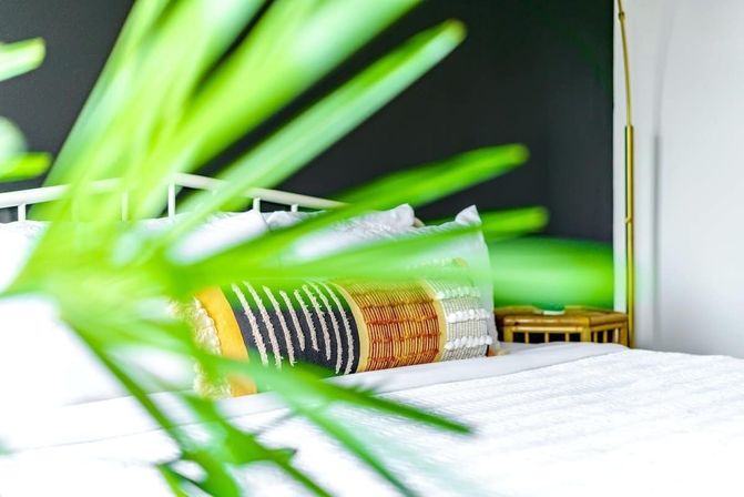 Bright modern bedroom with white bedding, a colorful patterned lumbar pillow, black accent wall, gold floor lamp and rattan bedside table, framed by blurred tropical plant leaves in the foreground.