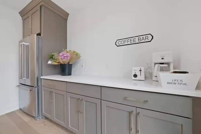 Modern kitchen coffee bar corner with stainless-steel French-door refrigerator, gray shaker cabinets, white countertop, coffee maker under a 'COFFEE BAR' sign, decorative tray reading 'LIFE IS BREW-TIFUL,' and a vase of flowers.