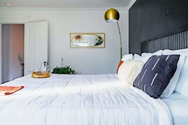 Bright modern bedroom interior with white comforter, dark gray accent wall, gold arched floor lamp and oval gold mirror, decorative pillows, plant and a tray with carafe on the bed.