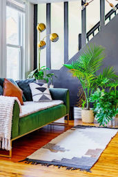 Bright modern living room with a green velvet sofa, geometric throw pillows and a cozy white blanket, gold triple-head floor lamp, potted tropical plants by a dark staircase, and a patterned runner rug on warm hardwood floors.