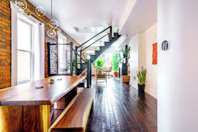Bright urban loft dining and living area with exposed brick walls, large windows, live-edge wooden dining table and bench, black metal open staircase, dark hardwood floors, globe pendant lights and abundant indoor plants