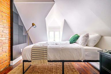 Cozy attic loft bedroom with sloped white ceilings and dormer window, white bed with green accent pillow and patterned throw, brass floor lamp, exposed brick wall and woven rug on hardwood floors.
