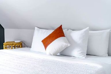 Bright minimalist bedroom with crisp white bedding, three white pillows and a brown-orange accent cushion on the bed, rattan bedside table and sloped white ceiling.
