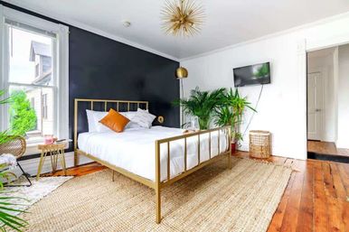 Sunlit modern bedroom with a gold metal bed frame, white bedding and orange accent pillow against a black accent wall, large window, potted green plants, woven jute rug and warm hardwood floors, minimalist boho style.