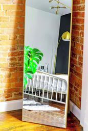 Full-length mirror reflecting a bright urban loft bedroom: white metal bed with quilt, exposed brick walls, hardwood floors, large monstera plant, and a gold arc floor lamp.