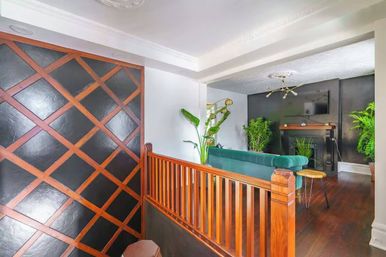 Cozy upstairs living room with green velvet sofa facing a black fireplace and wall-mounted TV, dark hardwood floors, warm wooden railing and diagonal lattice-accented black wall, brass chandelier and lush potted plants.