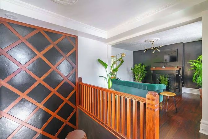 Cozy upstairs living room with green velvet sofa facing a black fireplace and wall-mounted TV, dark hardwood floors, warm wooden railing and diagonal lattice-accented black wall, brass chandelier and lush potted plants.
