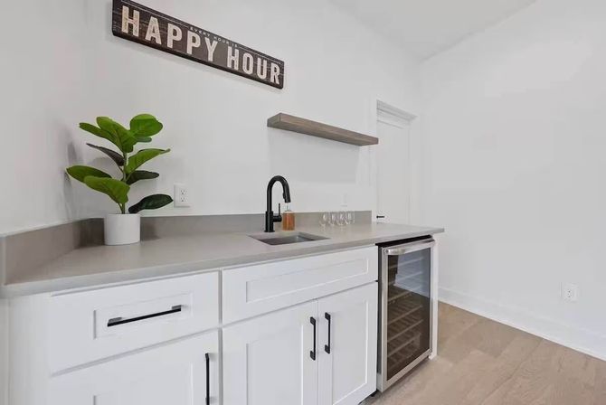 Bright modern wet bar/kitchenette with white cabinets, gray countertop, black faucet over a small sink, glass-front wine fridge, floating wood shelf, potted plant and a 'Happy Hour' wall sign.