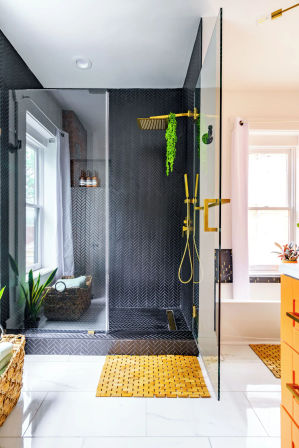 Stylish modern bathroom with glass-enclosed black herringbone-tile shower, gold rain and handheld fixtures, white marble floor, woven bath mats and potted greenery.