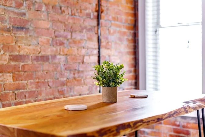 Cozy sunlit interior with an exposed brick wall and live-edge wooden table topped by a small potted green plant and two round coasters near a bright window.