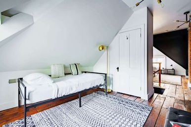 Cozy attic loft bedroom with sloped white ceiling, single black metal-frame bed dressed in white linens, patterned gray area rug on reclaimed hardwood floors, brass floor lamp and white closet door