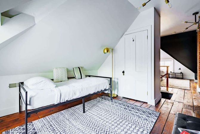 Cozy attic loft bedroom with sloped white ceiling, single black metal-frame bed dressed in white linens, patterned gray area rug on reclaimed hardwood floors, brass floor lamp and white closet door