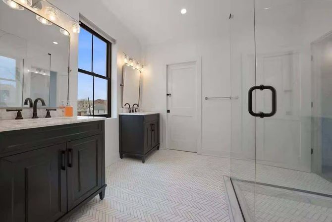 Bright modern bathroom with two black vanities and marble countertops, large black-framed window, glass-enclosed shower with matte-black hardware, and white herringbone tile floor.