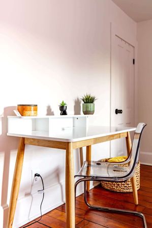 Bright minimalist home office with white mid-century desk and wooden legs, clear acrylic chair, potted succulents and woven basket on hardwood floor next to a white door.