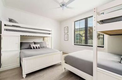 Bright sunlit modern bedroom with matching white bunk beds (twin-over-queen and twin), gray bedding, ceiling fan, wood-look floors, and a large window with blinds.