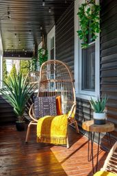 Sunlit covered front porch with a wicker egg chair, patterned pillow and mustard throw, hanging greenery, string lights, wooden deck and potted succulents
