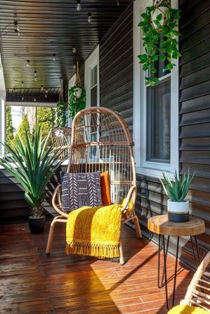 Sunlit covered front porch with a wicker egg chair, patterned pillow and mustard throw, hanging greenery, string lights, wooden deck and potted succulents